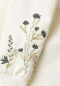 Tejido crema con intrincados bordados florales en azul oscuro y verde, con hojas en beige, dispuestos a lo largo de la manga.