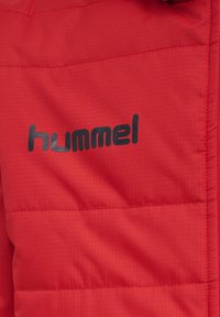 Rød pufferjakke med en quiltreret tekstur; har et sort "hummel" logo på fronten. Materialet ser letvægts og holdbart ud.