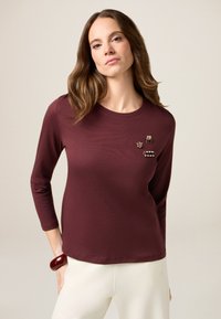 Burgunderfarbenes langärmliges Shirt mit rundem Halsausschnitt, verziert mit dekorativen Broschen. Weicher Stoff, lockere Passform und glatte Oberfläche.
