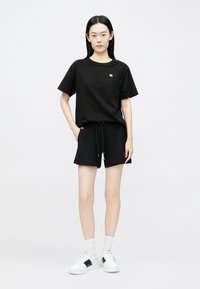 Fekete pamut póló kerek nyakkivágással, fekete rövidnadrággal, amely húzózsinóros derékrésszel rendelkezik, és fehér sportcipő fekete részletekkel.