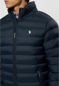Navy pufferjack met een hoge kraag, een horizontaal stikselpatroon, zijritsen en een klein geborduurd logo op de borst.