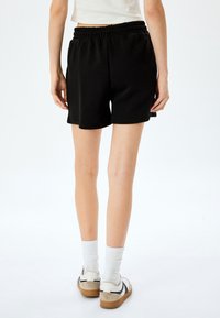 Pantalones cortos negros con cintura elástica, que llegan a mitad del muslo. Textura suave, diseño minimalista, combinados con calcetines blancos y zapatillas bajas.