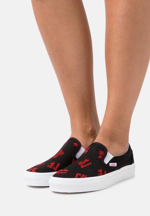 Jambes portant des baskets Vans slip-on noires à semelles blanches avec un motif de texte rouge "Waffle Lovers" sur un fond blanc uni.