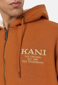 Sudadera con cremallera en color naranja, con un forro interior suave, que presenta bordado en hilo dorado de "KANI" y detalles de su establecimiento. Incluye capucha con cordón.