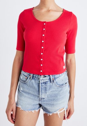 Femme portant un haut rouge à manches courtes côtelé boutonné, associé à un short en jean bleu clair usé.