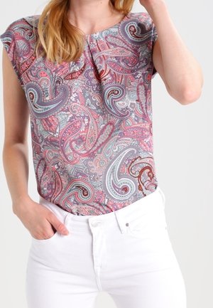 Femme portant un haut sans manches avec des motifs cachemire roses, violets et gris, rentré dans un pantalon blanc taille haute.