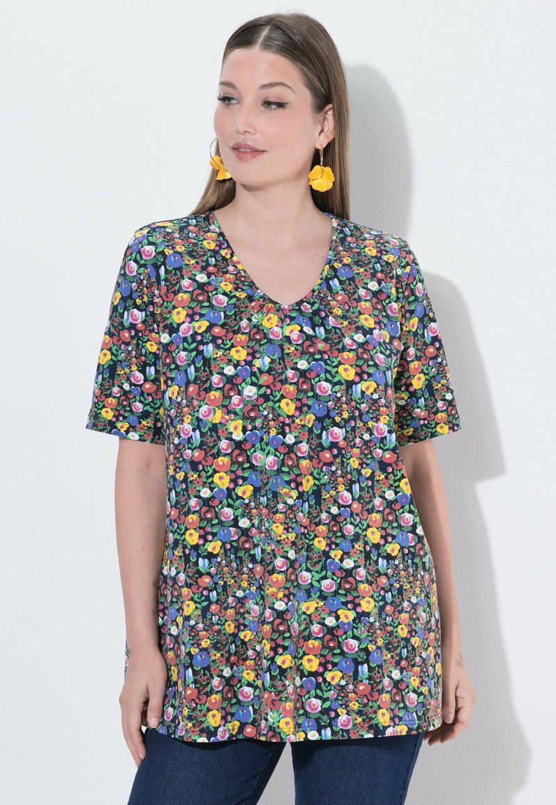 Kobieta nosząca żółte kolczyki w kształcie kwiatów oraz krótki top z dekoltem w serek i kolorowym kwiatowym wzorem, stojąca na białym tle.