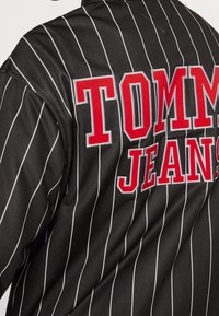 Svart satinjacka med tunna vita vertikala ränder. Har röd broderad text "TOMMY JEANS" på baksidan. Slät yta, rundad krage.