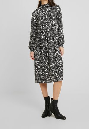 Žena v černobílých midi šatech s dlouhým rukávem a listovým vzorem s vysokým výstřihem a černými kotníkovými botami na podpatku.