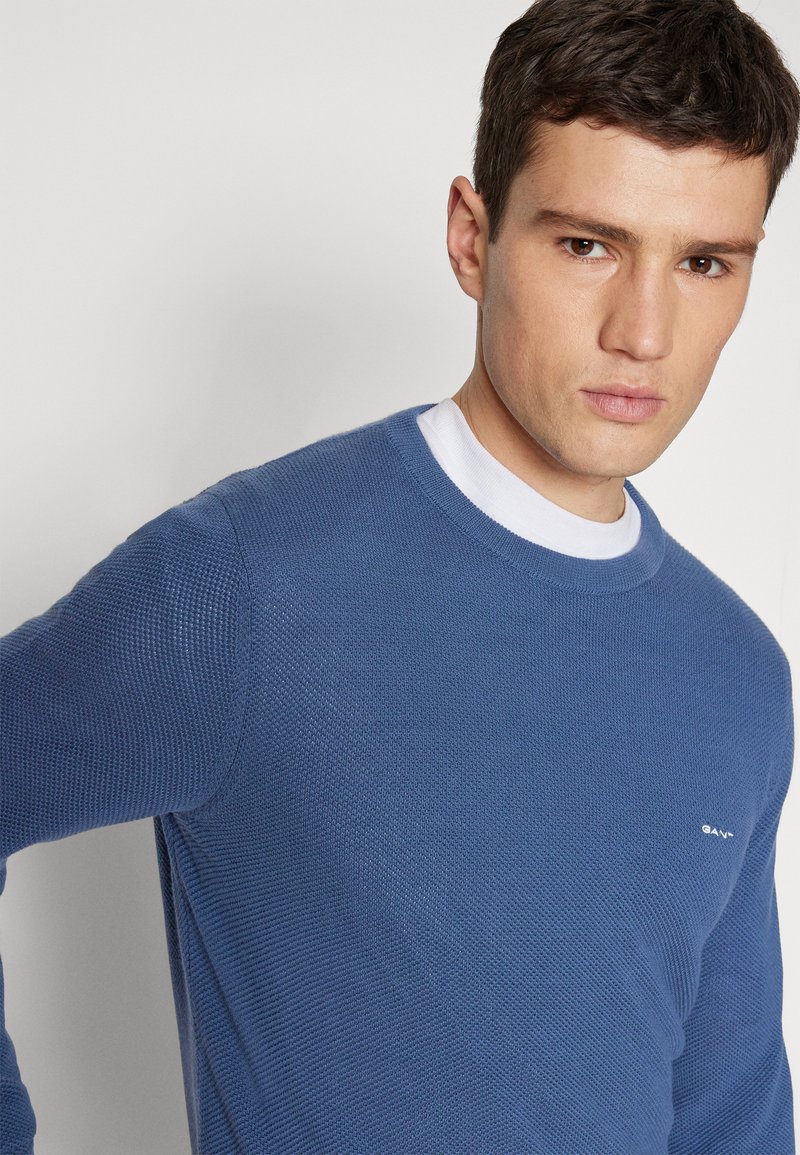 Suéter de punto azul con cuello redondo, tejido texturizado y logo blanco en el lado izquierdo del pecho. Capa interior suave visible en el cuello.