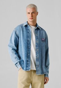 Ung mann med kort lyst hår iført en lyseblå denimskjorte, hvit T-skjorte og beige bukser, står foran en ensfarget grå bakgrunn.