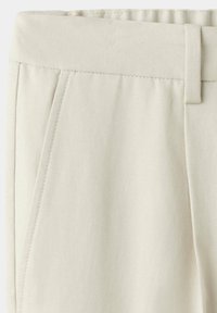 Gros plan sur la ceinture d’un pantalon en tissu beige avec passants pour ceinture et détails de couture de poche latérale.