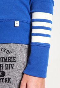 Sudadera azul con puños acanalados que presenta tres rayas blancas horizontales. Tela gris debajo con un gráfico de texto en negro. Diseño suave y casual.