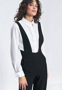 Czarny kombinezon z szerokimi ramiączkami na białej koszuli z kołnierzykiem. Posiada dopasowany krój i gładki materiał. Białe mankiety widoczne na nadgarstkach.