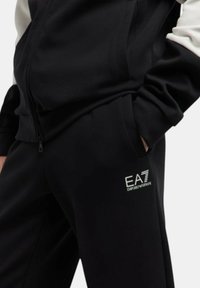 Completo tuta nera con giacca con zip e pantaloni. Presenta il logo "EA7 Emporio Armani" in bianco, con colori contrastanti nero e bianco, e una texture morbida.