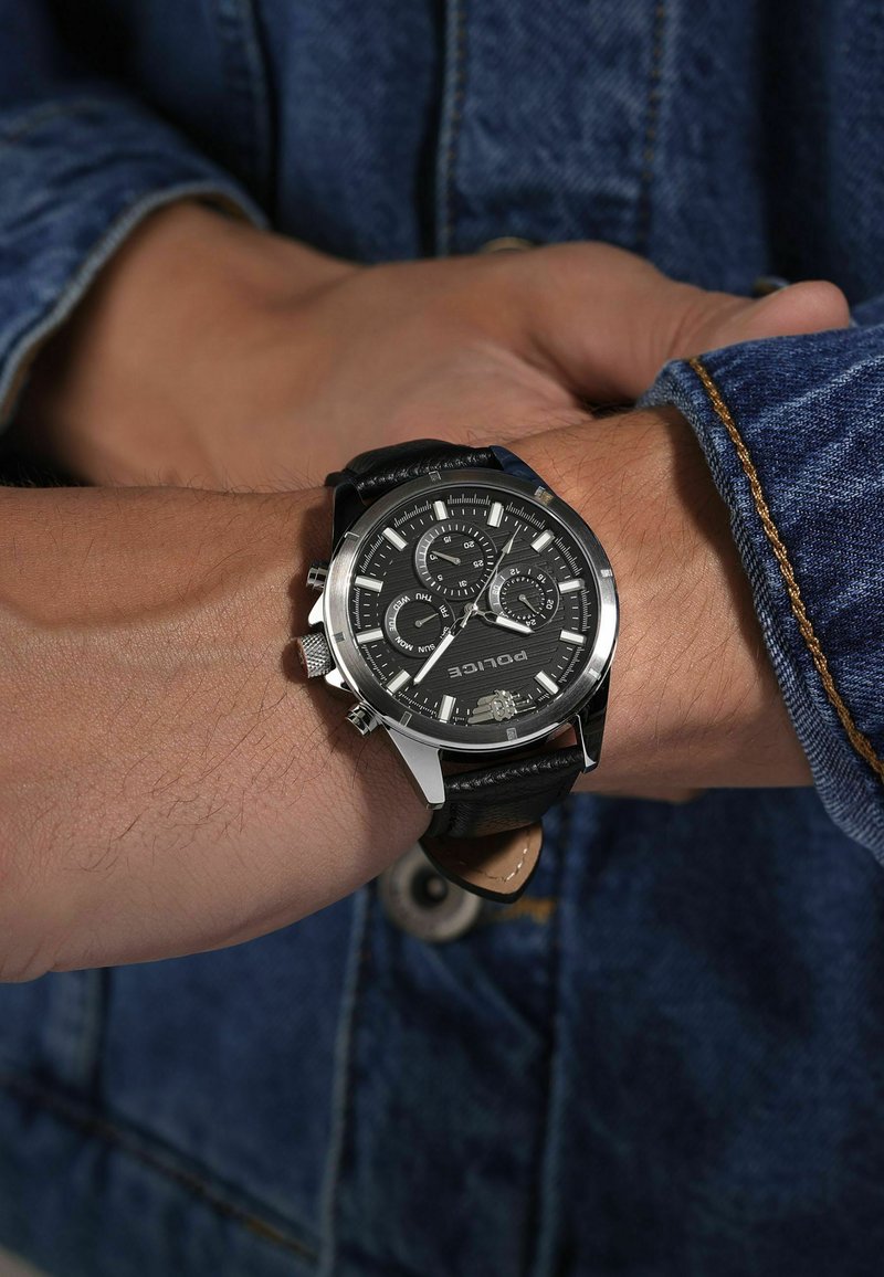 Montre chronographe noire avec accents argentés, cadran texturé et bracelet en cuir, portée sur un poignet sur fond en denim.
