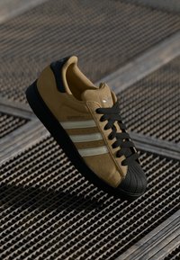 Yellow suede sneakers with black rubber toe cap, black laces, and three white stripes. Textured sole with light grip pattern.