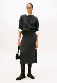 Jersey gris oscuro con rayas horizontales, camisa blanca con cuello debajo, falda larga a juego y botas negras hasta la rodilla; sostiene un bolso pequeño.