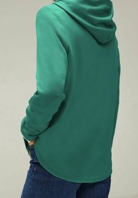 Zelená mikina s kapucí s texturovaným povrchem, vybavená stahovací šňůrkou v kapuci a dlouhými rukávy. Kombinována s modrým denimem, design je neformální a pohodový.