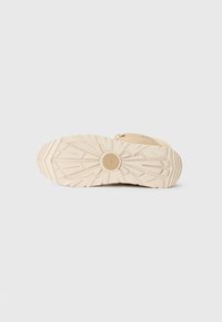 Suela de zapatilla en goma beige clara con un patrón de textura. Presenta un logo redondo en el centro y un diseño elevado para mayor tracción.