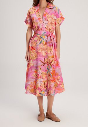 Robe portefeuille fleurie à manches courtes dans des nuances de rose et d’orange, nouée à la taille, portée avec des chaussures plates beiges.