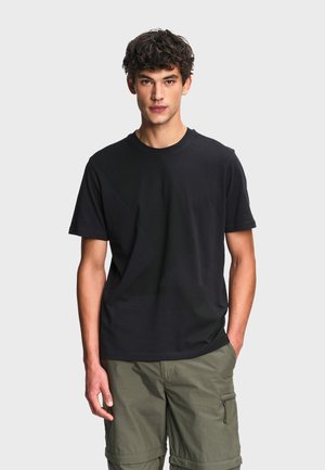 Jeune homme aux cheveux bouclés portant un t-shirt noir uni et un short cargo vert olive, debout devant un fond clair uni.