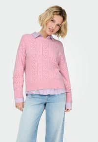 Pull rose tricoté avec motifs de cœurs, poignets côtelés, porté sur une chemise à col rayée bleu clair et blanc, assorti à un jean bleu clair.