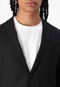 Sort blazer med en enkelt knap, slag og en brystlomme, lagdelt over en hvid t-shirt. Glat stofstruktur.
