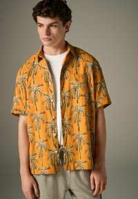Kurzarmhemd aus leuchtend orangefarbenem Stoff mit einem Muster aus Palmen in Schwarz und Grün. Mit Knöpfen vorne, lässige Passform.
