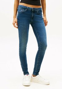 Jeans de mezclilla azul de corte slim con una cintura de altura media, diseño estándar de cinco bolsillos, ligero desgastado y detalles de costura sutiles. Combinados con zapatillas blancas.