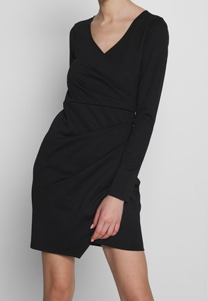 Kvinde iført en tætsiddende sort kjole med lange ærmer, slå-om front og V-udskæring, stående foran en ensartet grå baggrund.