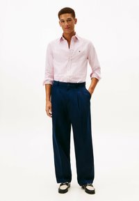 Camicia a righe rosa con bottoni abbinata a pantaloni blu navy plissettati e scarpe bicolore. La camicia ha un colletto classico e maniche arrotolate.