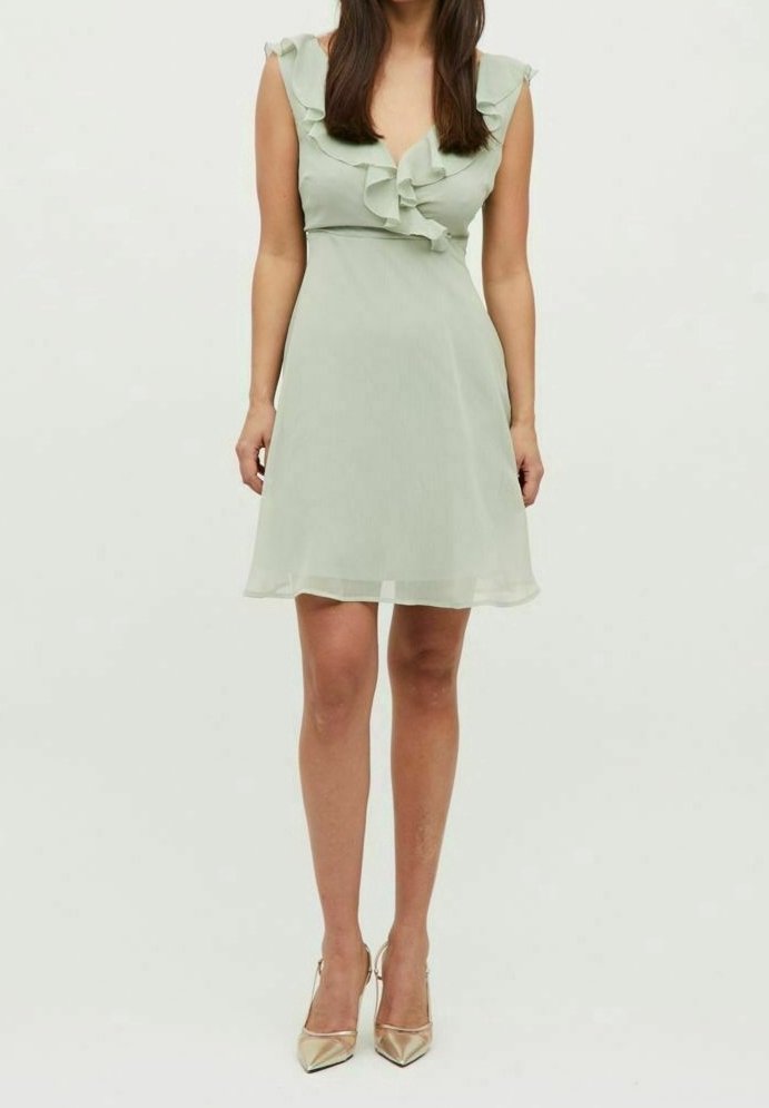 Femme portant une robe sans manches vert clair avec des volants et des talons pointus dorés, debout devant un fond blanc uni.
