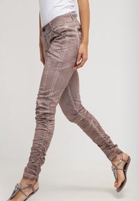 Jasnobrązowe obcisłe dżinsy o lekko pomarszczonej fakturze, z kieszeniami na zamek i detalami przeszyć, zestawione ze srebrnymi sandałami.