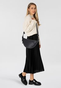 Borsa a tracolla in pelle nera con forma rotonda, texture liscia, tracolla regolabile e hardware in argento. Indossata con un maglione crema e una gonna plissettata.