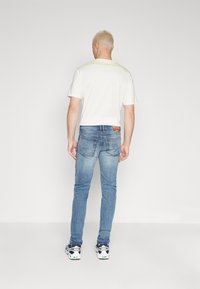 Vit t-shirt med kort ärm och neongul text på ryggen samt blå denimjeans. Jeansen har en smal passform och två bakfickor.