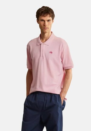 Uomo che indossa una polo rosa chiaro a maniche corte con piccolo logo sul petto e pantaloni casual blu scuro, mani in tasca, sfondo neutro.