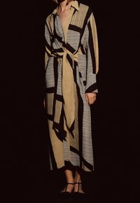 Frau in langem beige-schwarz gestreiften Kleid mit taillierter Taille und schwarzen T-Sandalen, die vor einem dunklen Hintergrund steht.