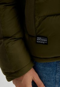 Chaqueta acolchada verde oliva con puños acanalados texturizados y una etiqueta en el costado que dice "INSIDE WORLDWIDE." Material suave y ligero.