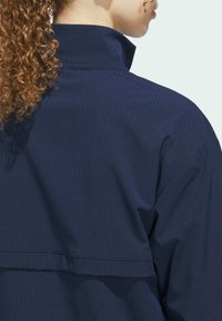 Navy blue jacket with a high collar, featuring a textured, ribbed pattern and layered back design. Visible seam detail enhances structure.