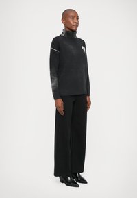 Sweater preto com colarinho alto em ribanas, apresentando uma textura em gradiente nas mangas e um pequeno design de coração. Combinado com calças largas pretas e botins.