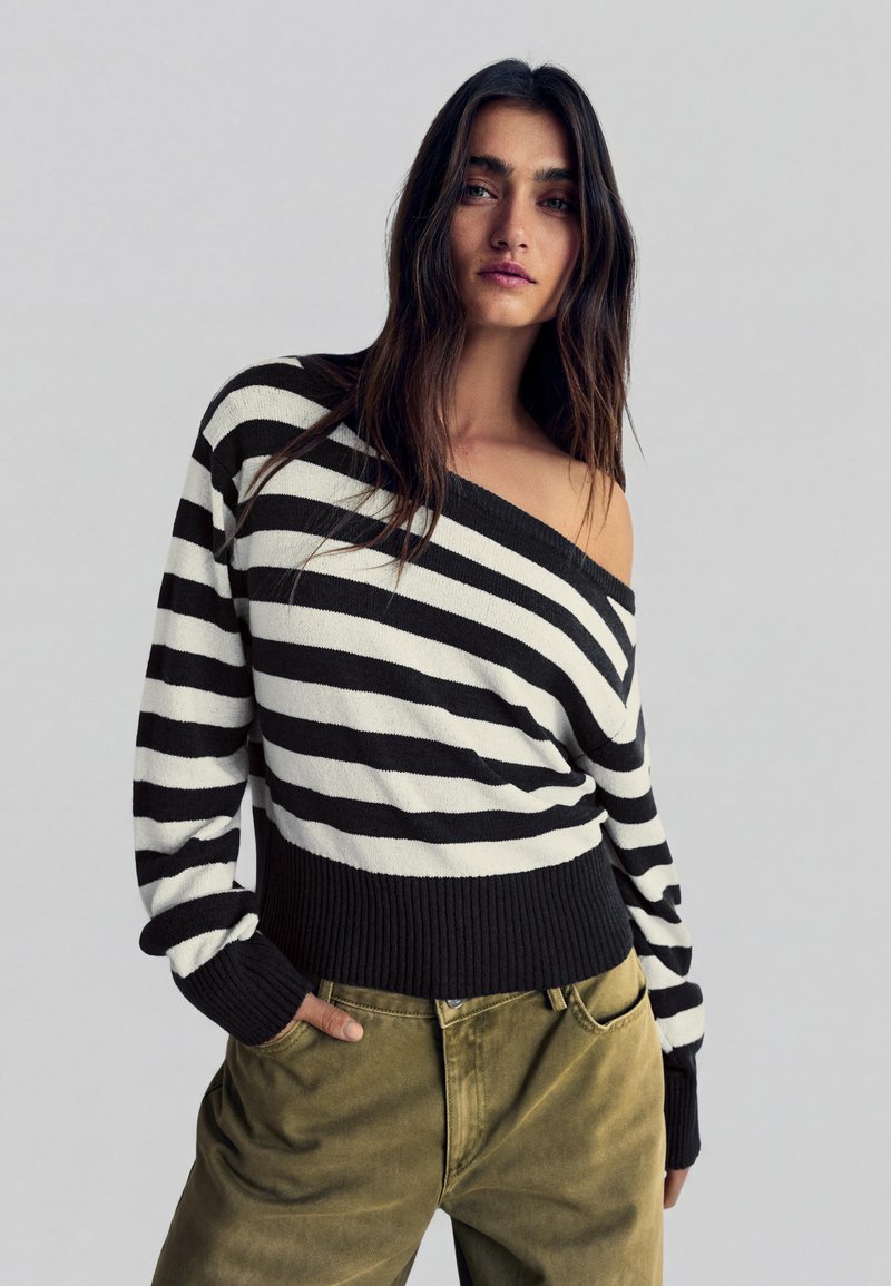 Femme aux longs cheveux foncés portant un pull rayé noir et blanc à épaules dénudées et un pantalon vert olive, les mains dans les poches.