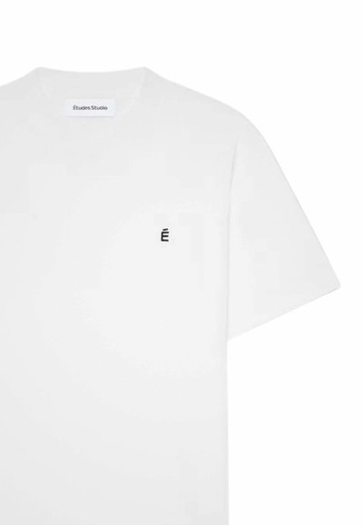 T-shirt blanc à manches courtes avec col rond, petit « É » noir sur la poitrine gauche et étiquette « Études Studio » à l'intérieur du col.
