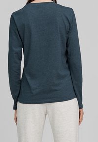 Kvinna iklädd en figursydd långärmad mörk turkos tröja och ljusa beige byxor, ståendes med ryggen mot kameran.