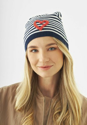Junge Frau mit langem blondem Haar, die eine marineblaue und weiße gestreifte Mütze mit einem roten Herzmotiv trägt, leicht lächelnd vor einem einfarbigen Hintergrund.