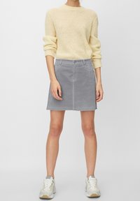 Falda de pana gris con un dobladillo ligeramente deshilachado, bolsillos frontales y cierre de botones, combinada con un suéter de punto de color beige claro y zapatillas.
