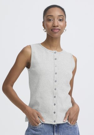 Femme portant un haut gris sans manches boutonné et un jean bleu, mains dans les poches, fond neutre.