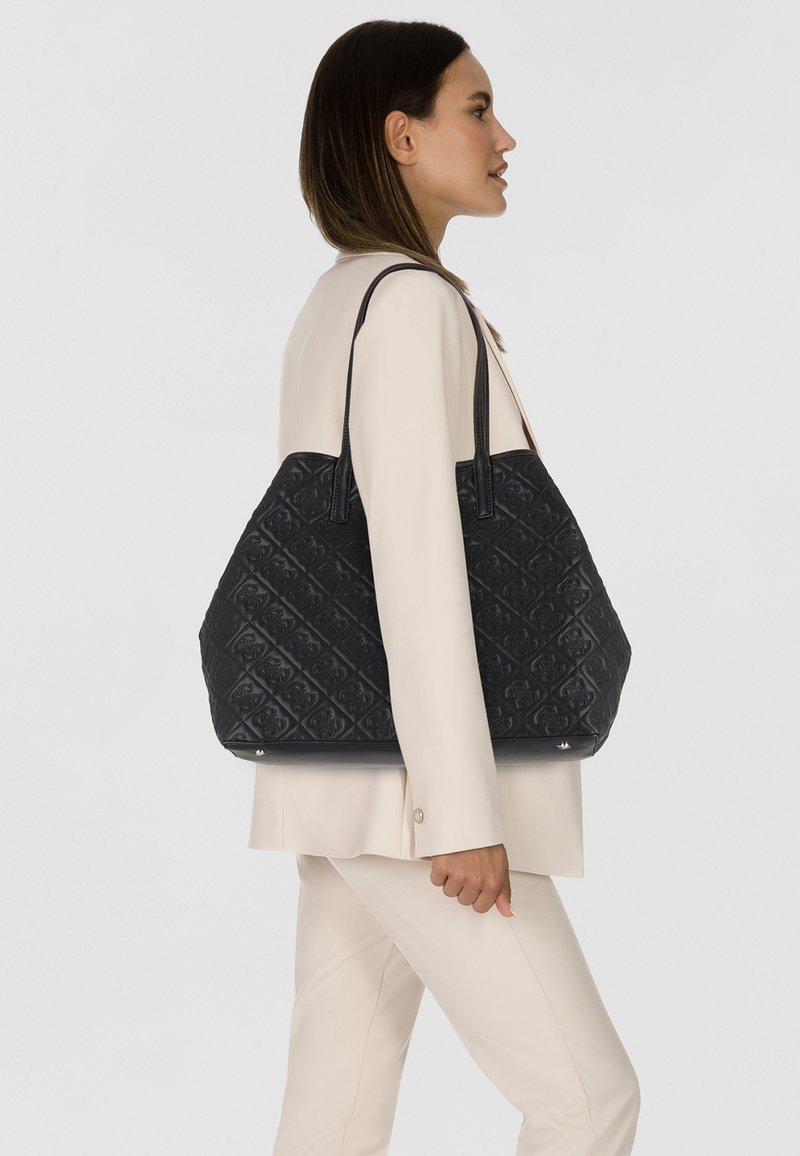 Sac fourre-tout noir matelassé avec un motif floral embossé, des poignées en cuir et une base plate. Ce sac présente un design spacieux et structuré.
