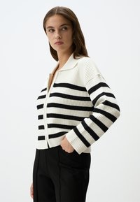 Cardigan lavorato a maglia di colore crema con strisce orizzontali nere, con zip sul davanti e colletto a punta. Texture a costine con maniche larghe.