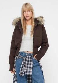 Mladá žena s dlouhými blond vlasy, oblečená v tmavě hnědé péřové bundě s kapucí podšitou kožešinou, světle šedém crop topu, modrých džínech a zavázané kostkované košili.